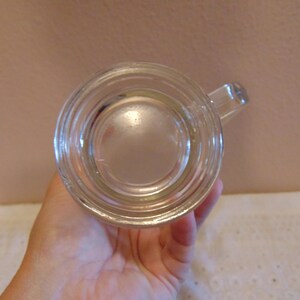 May include: A clear glass mug viewed from directly above, showing a circular base with concentric ring indentations and a small handle extending from the side. The glass is transparent, revealing the smooth, reflective surface of the bottom.