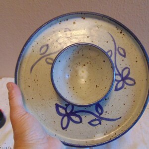 May include: A round ceramic serving dish with a central bowl, featuring a speckled off-white glaze and hand-painted dark blue floral motifs. The rim is finished with a dark blue band, and the piece is shown next to a black measuring tape.