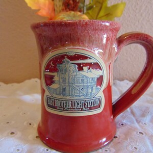 May include: A red ceramic mug with a glossy finish and a large handle. The front features an oval decal depicting the East Brother Light Station in San Francisco Bay, with the text 1873, East Brother Light Station, and San Francisco Bay.