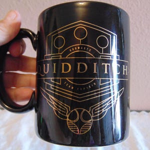 May include: Black ceramic mug with gold lettering and design. The mug features the word "QUIDDITCH" and the words "Hogwarts Team Captain". The design includes wings and a golden snitch.