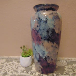 May include: A ceramic vase with a mottled, multi-colored glaze featuring shades of deep purple, light blue, soft pink, and cream. The vase has a cylindrical body that tapers slightly toward the base and a wide, rounded rim at the top opening.
