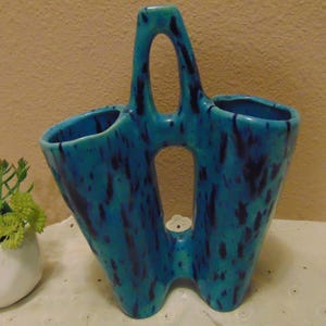 May include: A dual-chambered ceramic vase featuring a vibrant turquoise glaze with dark blue vertical splatters. The piece has a central handle with two open cutouts, creating a distinct silhouette with two separate openings for floral arrangements.