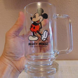 May include: A clear glass mug featuring a graphic of Mickey Mouse wearing red shorts and yellow shoes. The text MICKEY MOUSE is printed in black below the character, followed by the smaller text &copy;WALT DISNEY PRODUCTIONS. The mug has a handle.