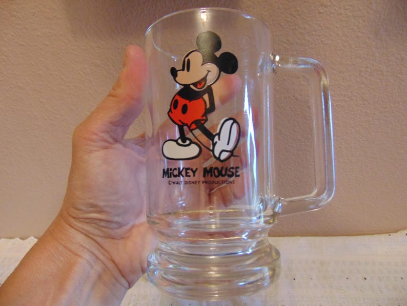 May include: A clear glass mug featuring a graphic of Mickey Mouse wearing red shorts and yellow shoes. The text MICKEY MOUSE is printed in black below the character, followed by the smaller text &copy;WALT DISNEY PRODUCTIONS. The mug has a handle.