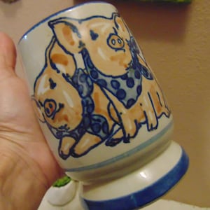 May include: A ceramic mug with a cream-colored base featuring a hand-painted illustration of two light orange pigs wearing blue polka-dot bandanas. The mug has a flared base and a blue rim, with blue painted accents around the bottom and top edges.