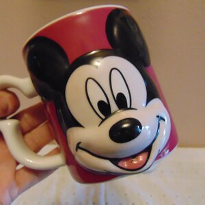 May include: A ceramic mug featuring a three-dimensional molded face of Mickey Mouse on the front. The mug is primarily red with black ears and a white, black, and pink face design. It includes a white handle shaped like a stylized Mickey Mouse icon.