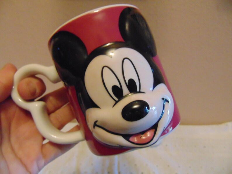 May include: A ceramic mug featuring a three-dimensional molded face of Mickey Mouse on the front. The mug is primarily red with black ears and a white, black, and pink face design. It includes a white handle shaped like a stylized Mickey Mouse icon.
