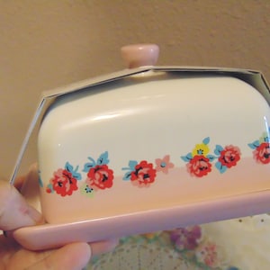 May include: A ceramic butter dish with a lid and base, featuring a floral design. The dish is primarily white with a pink ombre effect at the base. Red, blue, and yellow flowers decorate the front.
