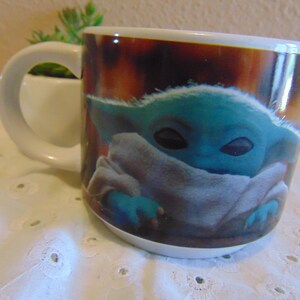 May include: A white ceramic mug featuring a printed graphic of a small, teal-skinned creature with large dark eyes and pointed ears, wrapped in a light-colored fabric. The mug has a curved handle and a smooth, glossy finish on the exterior surface.