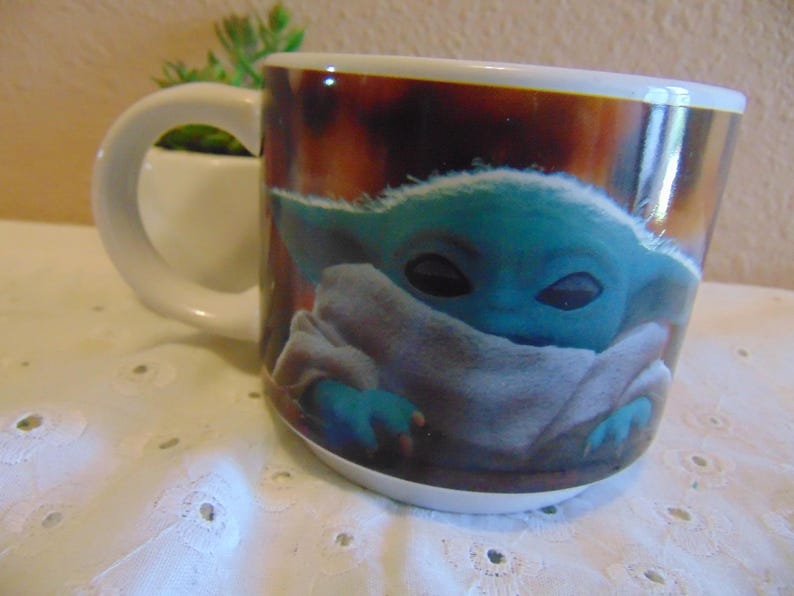 May include: A white ceramic mug featuring a printed graphic of a small, teal-skinned creature with large dark eyes and pointed ears, wrapped in a light-colored fabric. The mug has a curved handle and a smooth, glossy finish on the exterior surface.