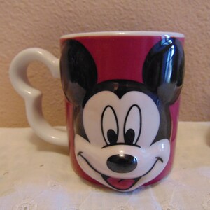 May include: A ceramic mug featuring a raised, three-dimensional molded face of Mickey Mouse on a deep pink background. The mug has a white handle shaped like a curved silhouette of Mickey Mouse ears and a smooth, glossy finish throughout the exterior.