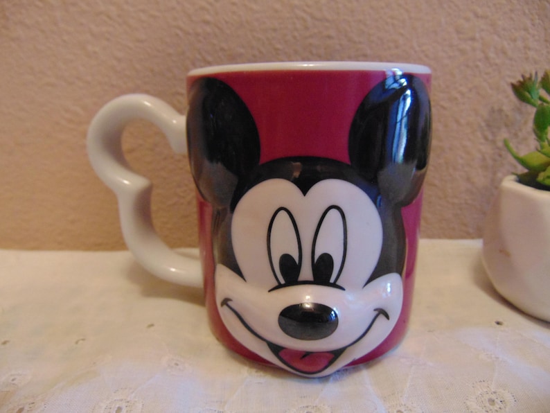 May include: A ceramic mug featuring a raised, three-dimensional molded face of Mickey Mouse on a deep pink background. The mug has a white handle shaped like a curved silhouette of Mickey Mouse ears and a smooth, glossy finish throughout the exterior.