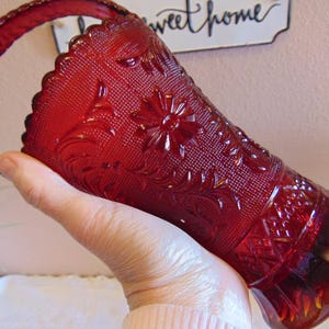 May include: A ruby red glass basket with a handle, featuring an embossed floral and geometric pattern. The basket has a scalloped rim and a textured surface. The handle is also textured and has a rounded shape. The words "sweet home" are visible in the background.