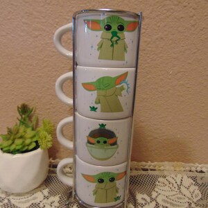 May include: A set of four white ceramic mugs stacked vertically in a chrome metal stand. Each mug features a green character illustration with large ears and a tan robe in various poses, including holding a frog and using a blue energy force.