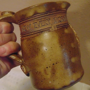 May include: A brown ceramic mug with a speckled finish and a handle. The upper portion of the mug features an embossed band with the text NEW EVERY MORN, which is partially visible. The mug has a cylindrical body with a slightly flared rim.