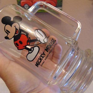 May include: A clear glass mug featuring a graphic of Mickey Mouse wearing red shorts and yellow shoes. The text MICKEY MOUSE and &copy;WALT DISNEY PRODUCTIONS is printed in black on the side of the glass. The mug has a sturdy handle and a thick base.