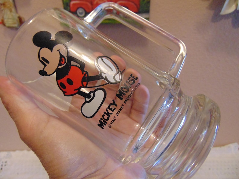 May include: A clear glass mug featuring a graphic of Mickey Mouse wearing red shorts and yellow shoes. The text MICKEY MOUSE and &copy;WALT DISNEY PRODUCTIONS is printed in black on the side of the glass. The mug has a sturdy handle and a thick base.