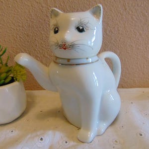 May include: A white ceramic teapot shaped like a sitting cat with a removable head lid. The cat features painted black eyes, whiskers, and a gold-trimmed collar with gold dots. A curved handle is on the back and a spout extends from the front.