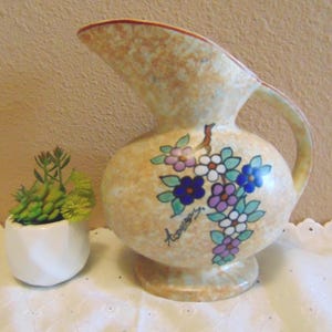 May include: A vintage, speckled beige pitcher with a floral design in blue, purple, and white. The pitcher has a handle and spout, and sits next to a small white pot with green succulents.