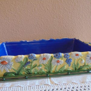 May include: A rectangular ceramic baking dish with a vibrant yellow exterior featuring raised, hand-painted white daisy flowers and green foliage. The interior is a solid deep blue. The dish rests on a dark green metal wire stand.
