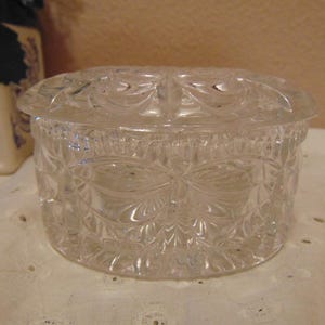 May include: A clear, round glass trinket box with a removable lid. The exterior of the container and the lid feature a pressed, faceted geometric pattern that creates a textured, reflective surface across the entire piece of glassware.
