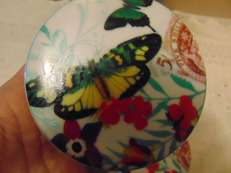 Pu&ograve; includere: Coperchio decorativo rotondo e bianco con un motivo a farfalla con ali gialle, verdi e nere. Fiori rossi e foglie verdi circondano la farfalla. Il coperchio presenta anche un design di francobollo vintage.