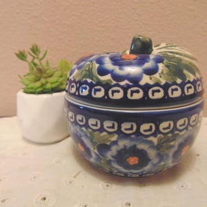 May include: A ceramic apple-shaped lidded jar featuring a hand-painted blue floral pattern with green leaves and orange centers. The design includes a repeating geometric border of blue and white squares around the center and lid rim.