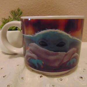 May include: A white ceramic mug featuring a printed graphic of a small, green, large-eared creature with dark eyes wrapped in a light-colored robe. The mug has a rounded handle on the left side and a smooth, glossy finish, showcasing a vibrant, warm-toned background behind the character illustration.