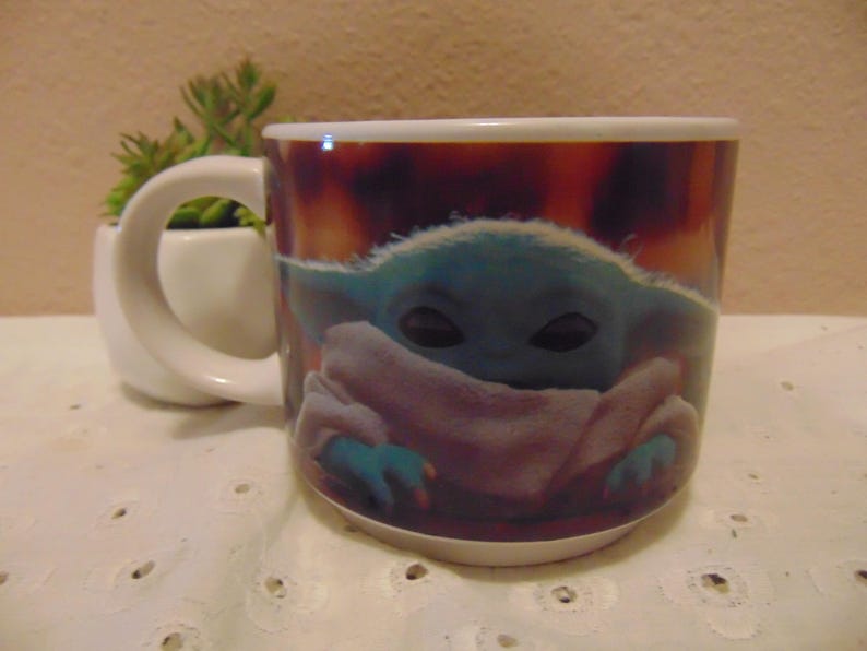 May include: A white ceramic mug featuring a printed graphic of a small, green, large-eared creature with dark eyes wrapped in a light-colored robe. The mug has a rounded handle on the left side and a smooth, glossy finish, showcasing a vibrant, warm-toned background behind the character illustration.