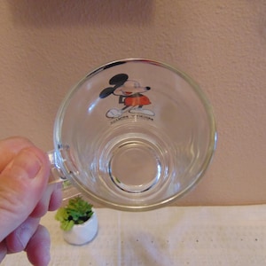 May include: A clear glass mug featuring a graphic of Mickey Mouse on the interior base. The character is depicted in black, red, and white colors. The text Mickey Mouse is printed in black lettering below the character figure on the glass surface.