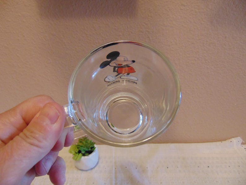 May include: A clear glass mug featuring a graphic of Mickey Mouse on the interior base. The character is depicted in black, red, and white colors. The text Mickey Mouse is printed in black lettering below the character figure on the glass surface.