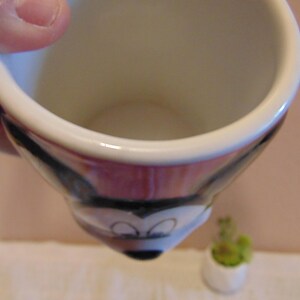 May include: A ceramic mug featuring a molded design of the face of Mickey Mouse. The mug has a white interior and a handle on the side. The exterior surface displays the black, white, and red colors characteristic of the character's face.