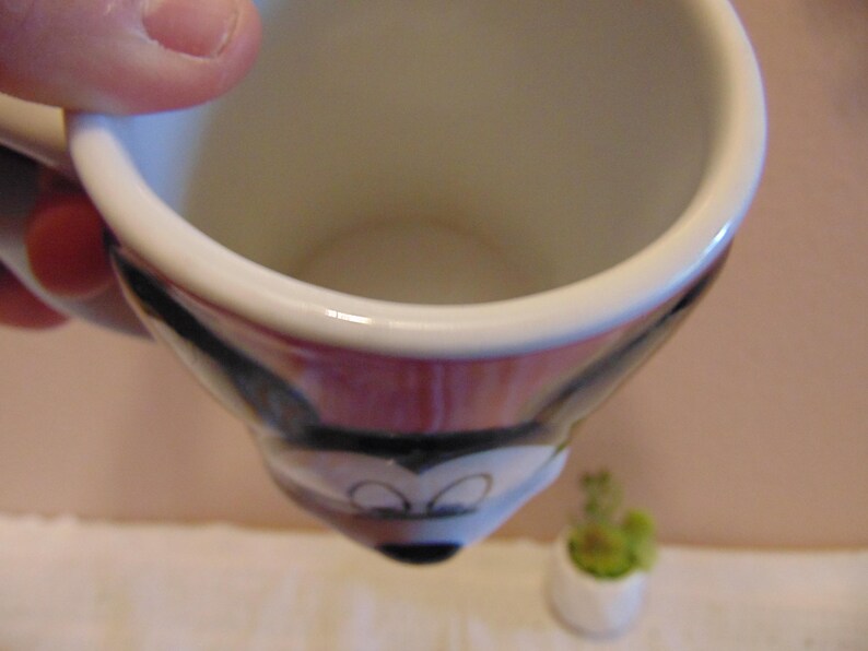 May include: A ceramic mug featuring a molded design of the face of Mickey Mouse. The mug has a white interior and a handle on the side. The exterior surface displays the black, white, and red colors characteristic of the character's face.
