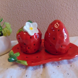 May include: A ceramic salt and pepper shaker set shaped like red strawberries with black seeds, featuring a white flower on one shaker. The shakers rest on a matching red strawberry-shaped tray with a green leaf accent on the side.