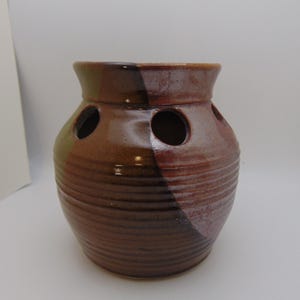 May include: A bulbous ceramic vase with a glossy brown glaze and horizontal ribbed texture. The vase features a wide neck and three circular openings spaced evenly around the upper shoulder, showcasing a blend of dark brown and earthy tones.