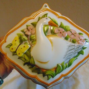 May include: A white ceramic wall hanging with a swan design. The swan is surrounded by pink flowers, green leaves, and yellow butterflies. The border is orange and the piece has a scalloped edge.