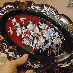 May include: An oval-shaped decorative tray featuring a deep red center with iridescent mother-of-pearl floral inlays. The wide, scalloped black border is accented with gold-colored flecks and additional silver-toned mother-of-pearl scrollwork.