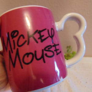 May include: A cylindrical ceramic mug with a vibrant pink exterior and a white interior. The front features the text Mickey Mouse in bold black lettering. The handle is shaped like the iconic three-circle silhouette of a mouse head.