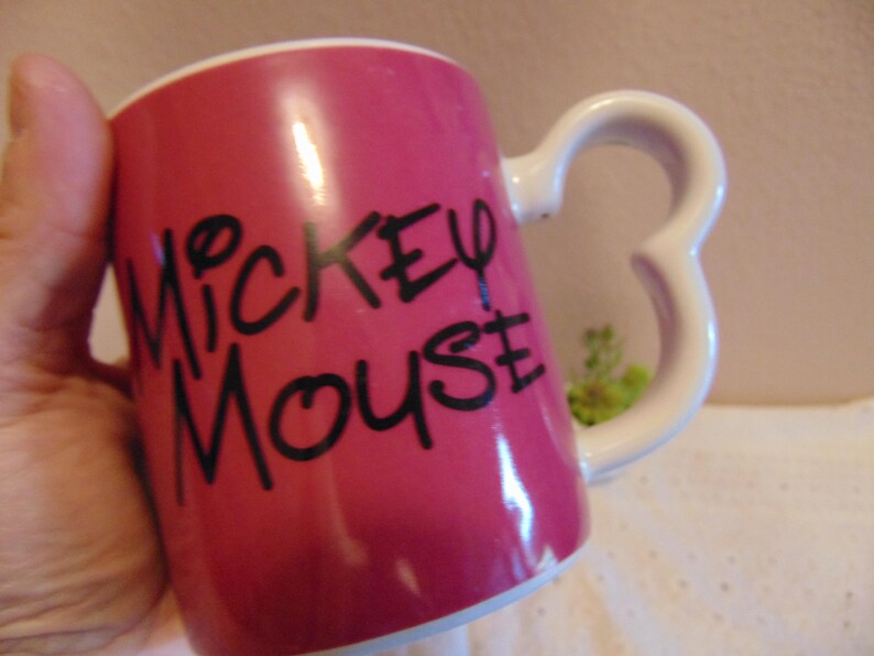 May include: A cylindrical ceramic mug with a vibrant pink exterior and a white interior. The front features the text Mickey Mouse in bold black lettering. The handle is shaped like the iconic three-circle silhouette of a mouse head.