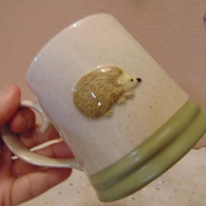 May include: A ceramic mug with a speckled off-white finish and a raised, three-dimensional brown hedgehog figure on the side. The base of the mug features a wide, solid sage green band, and it includes a curved handle on the left side.
