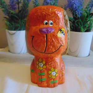 May include: Orange ceramic dog figurine with a smiling face, purple nose, and yellow flowers painted on its body. A small yellow and black butterfly is painted on the dog's cheek.