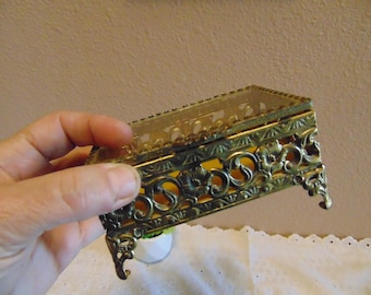 May include: An antique brass jewelry box with ornate filigree detailing. The rectangular box has a clear glass top and rests on four decorative feet. The brass has a warm, aged patina, and the interior is lined with a yellow material.