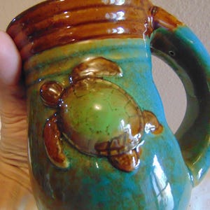 May include: A ceramic mug featuring a raised, three-dimensional sea turtle relief on the side. The mug has a glossy glaze with a blend of turquoise, teal, and brown colors, including a dark brown rim and a sturdy, curved handle on the right side.