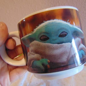 May include: A white ceramic mug featuring a printed graphic of a small, green, large-eared creature with dark eyes, wrapped in a beige textured robe. The background of the print is a warm, mottled brown color, and the mug has a simple handle.