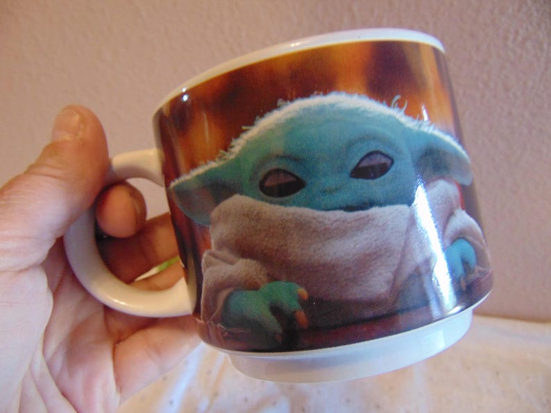 May include: A white ceramic mug featuring a printed graphic of a small, green, large-eared creature with dark eyes, wrapped in a beige textured robe. The background of the print is a warm, mottled brown color, and the mug has a simple handle.