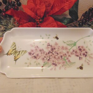 May include: A rectangular white ceramic tray with scalloped edges, featuring a delicate illustration of light purple lilac flowers, a yellow and black striped butterfly, and two small bees on the surface of the dish.
