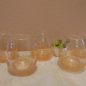 May include: A set of five clear stemless wine glasses, each featuring a rounded bowl shape. The base of every glass is decorated with a dense, speckled pattern of fine, shimmering orange glitter that fades toward the clear upper rim.