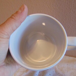 May include: A plain white ceramic mug with a smooth, glossy finish and a rounded handle. The interior of the mug is entirely white and empty, showing a clean, circular opening. The mug is designed with a simple, functional structure for holding liquids.