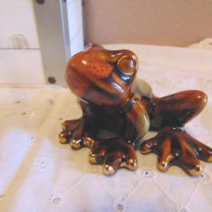 May include: A ceramic frog figurine in shades of brown and gold. The frog has large, expressive eyes and gold-tipped feet. The glossy finish gives the figurine a polished look, and it sits on a white, patterned surface.