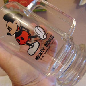 May include: A clear glass mug featuring a graphic of Mickey Mouse in red shorts and yellow shoes. The text MICKEY MOUSE and WALT DISNEY PRODUCTIONS is printed in black. The mug has a thick handle and a ribbed base design for a sturdy grip.